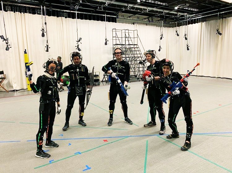 Jamie standing in the center of a mocap session, holding a plastic gun, surrounded by other actors